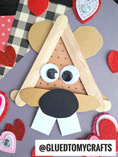 A popsicle stick groundhog face made from sticks, paper, and large googly eyes is surrounded by felt and paper hearts on a purple background, with the text @GLUEDTOMYCRAFTS at the bottom.