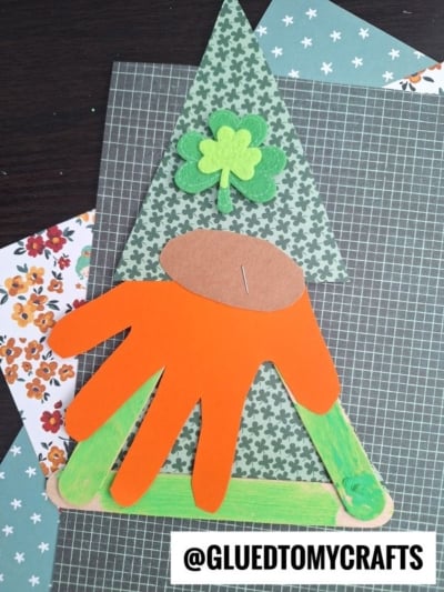 A Popsicle Stick Leprechaun Gnome craft for St. Patrick’s Day, featuring patterned paper, a green shamrock, an orange handprint beard, and green-painted sticks, displayed on patterned sheets. @GLUEDTOMYCRAFTS is at the bottom.