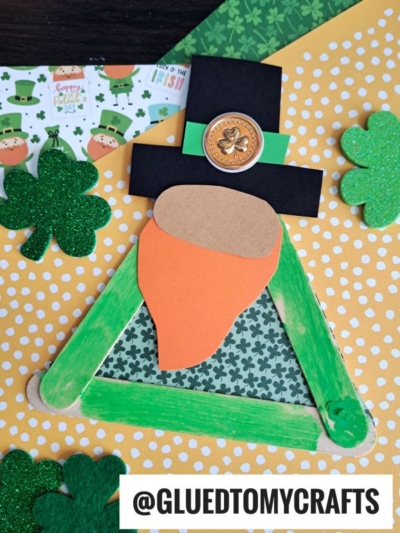 A St. Patrick’s Day Popsicle Stick Leprechaun Gnome craft featuring a whimsical gnome leprechaun face, paper beard, and shamrock hat. Surrounded by festive green-patterned papers. Text reads “@GLUEDTOMYCRAFTS”.