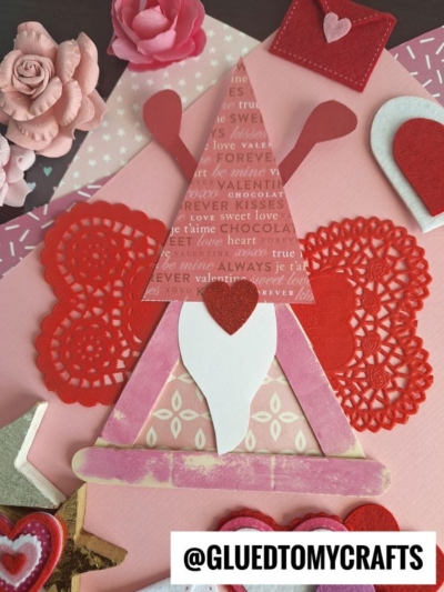 A Valentine gnome love bug craft made from red and pink paper, doilies, and a wooden stick, surrounded by heart and flower decorations. The Instagram handle @GLUEDTOMYCRAFTS is at the bottom.