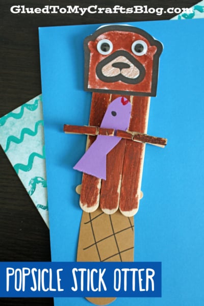 A charming popsicle stick otter craft featuring a brown otter face, googly eyes, and a purple fish held in its arms, displayed on a blue background with the text “POPSICLE STICK OTTER.”.
