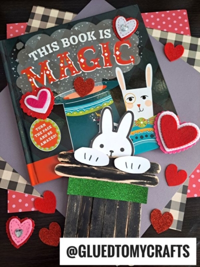 A children’s book titled “This Book Is Magic” is surrounded by red and pink felt hearts. In front of the book, a craft features a paper bunny popping out of a popsicle stick magician’s hat, with scrapbook paper as the background.