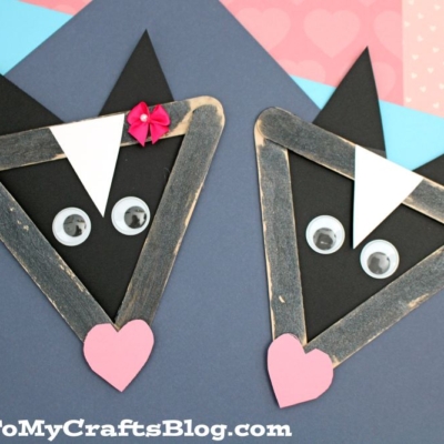 Two popsicle stick skunk crafts made from triangle-shaped sticks, black paper, googly eyes, and heart-shaped noses are displayed on colorful paper backgrounds. One skunk has a pink bow on its head.