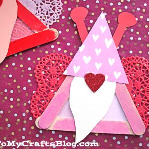 Two handmade valentine gnome crafts made from popsicle sticks, colored paper, and heart-shaped doilies on a pink polka dot background. The gnomes wear heart-themed hats and have white beards, perfect for a love bug craft session.
