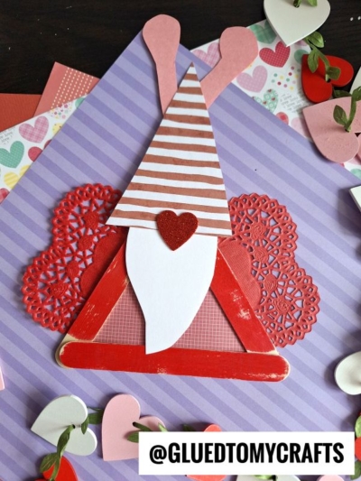 A Valentine gnome love bug craft made from paper, featuring a red triangle body, striped hat with heart antennae, white beard, and a red heart nose, set on purple striped paper with heart decorations all around.