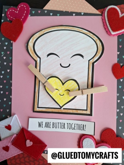 A handmade Punny Toast Card features a smiling bread slice holding a yellow heart with clothespins, surrounded by red felt hearts and mini felt envelopes. Text reads “WE ARE BUTTER TOGETHER!” and “@GLUEDTOMYCRAFTS.”.