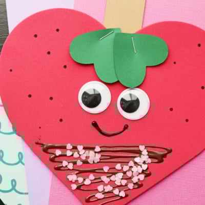 A handmade Valentine’s Day card shaped like a heart shaped strawberry with googly eyes, a smiling mouth, green paper leaves, and brown chocolate details with pink heart sprinkles. The card is on pastel colored paper.