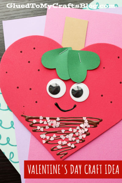 A handmade Valentine’s Day card shaped like a heart shaped strawberry with googly eyes, a smiling mouth, green paper leaves, and brown chocolate details with pink heart sprinkles. The card is on pastel colored paper.