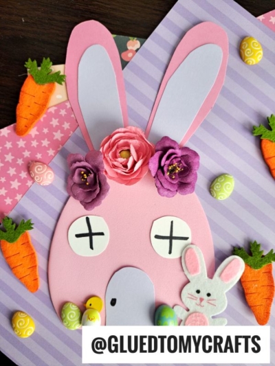 A pink paper bunny face with flowers on its head, closed eyes marked by Xs, and surrounded by felt carrots, mini eggs, and a small foam egg house on a striped and polka-dot background. @GLUEDTOMYCRAFTS text at the bottom.