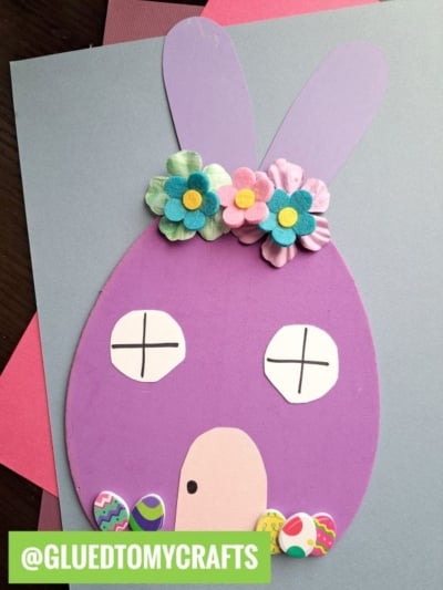 A purple bunny-shaped craft form bunny house with two felt flowers on its head, simple white eyes with X marks, a tan oval nose, and decorative Easter egg stickers near its mouth. @GLUEDTOMYCRAFTS text label at the bottom left.