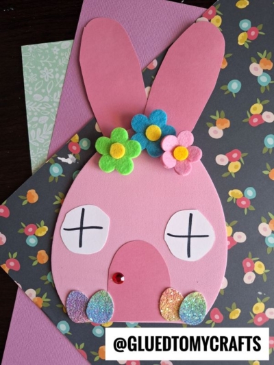 A pink bunny face made from paper with closed eyes, colorful flower decorations, and glittery egg shapes at the bottom creates the perfect Easter bunny house craft. The floral patterned background adds charm. Text reads @GLUEDTOMYCRAFTS.