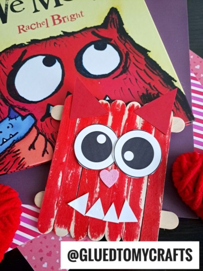 A red craft stick love monster with big googly eyes, pointy ears, and white teeth sits next to a children's book featuring a similar monster. @GLUEDTOMYCRAFTS is at the bottom.