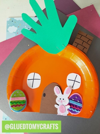 A craft project featuring an orange paper plate carrot house decorated with a green handprint roof, paper windows, brown door and chimney, two felt Easter eggs, and a felt bunny on a colorful background.