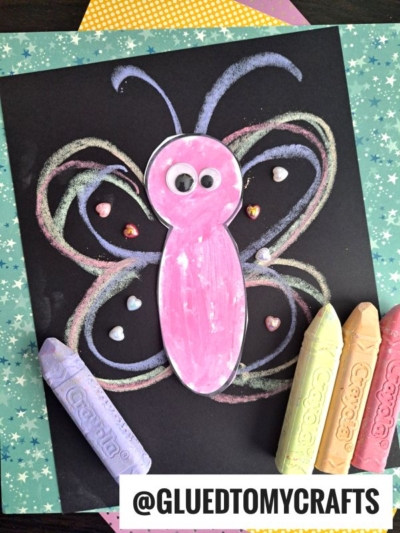 A pink paper butterfly with googly eyes is glued to black paper. Colorful chalk outlines form the wings in this creative chalk butterfly craft, with small heart-shaped gems as decoration. Four pastel crayons are placed beside the artwork.