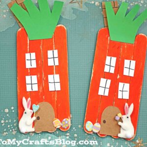 Two popsicle stick carrot houses, crafted from orange sticks with green paper tops and white paper windows, are decorated with bunny and chick stickers. They sit on a blue background sprinkled with stars—an adorable popsicle stick carrot house scene.
