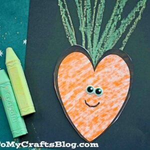 A heart-shaped orange carrot with googly eyes and a smile is glued to black paper.