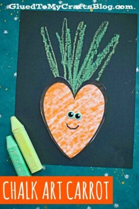 A heart-shaped orange carrot with googly eyes and a smile is glued to black paper. Green crayon strokes top the chalk art carrot, and two crayons lie on the side. Text reads Chalk Art Carrot.