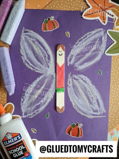 A butterfly craft with a popsicle stick body and chalk art fairy wings on purple paper, surrounded by stickers, crayons, and school glue. The butterfly has a smiling face and a green pattern on the wings.
