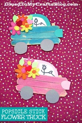 Two colorful popsicle stick spring flower trucks, one blue and one pink, decorated with paper flowers and gray wheels, are displayed on a pink polka dot background. Each truck features a smiling stick figure driver.