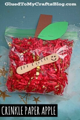 A crinkle paper apple craft: a ziplock bag filled with red and gold crinkle paper, decorated with a green paper leaf, brown paper stem, and a smiling craft stick worm. Text reads CRINKLE PAPER APPLE.
