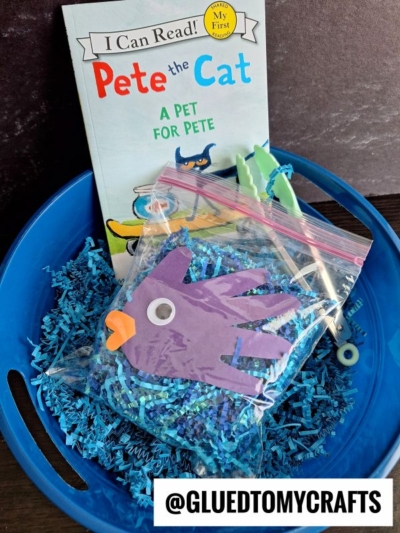 A blue basket with crinkle paper holds a Pete the Cat: A Pet for Pete book and a bag featuring a purple handprint paper fish craft, googly eye, and clothespin—perfect for any crinkle paper fish bowl project. Watermarked @GLUEDTOMYCRAFTS.