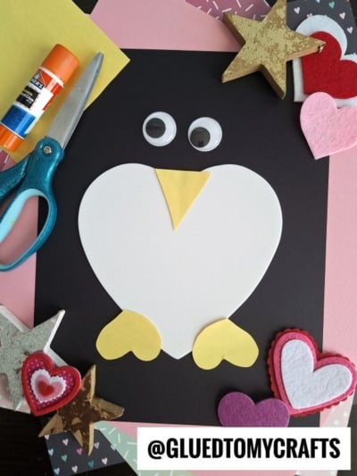 A penguin heart valentine craft made with cut-out shapes on black paper, surrounded by scissors, glue, felt hearts, and sparkly stars. The penguin has googly eyes, yellow feet, a yellow beak, and a sweet heart theme. @GLUEDTOMYCRAFTS appears at the bottom.