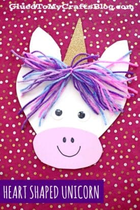 A colorful unicorn craft made from heart-shaped paper pieces, yarn hair, and a gold horn, perfect for heart shaped crafts or valentine art project ideas. Set against a red polka dot background. Text reads HEART SHAPED UNICORN.