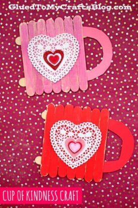 Two handmade popsicle stick cup of kindness crafts, decorated with doilies and hearts, are displayed on a pink polka dot background. One charming cup is pink, while the other is red.