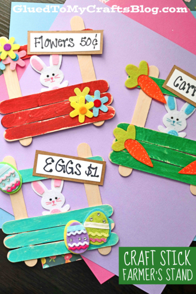 Three colorful Popsicle Stick Farmer's Stand signs shaped like stands, decorated with paper bunnies, flowers, carrots, and eggs. Each sign shows handwritten prices for flowers, carrots, and eggs on a pink background.