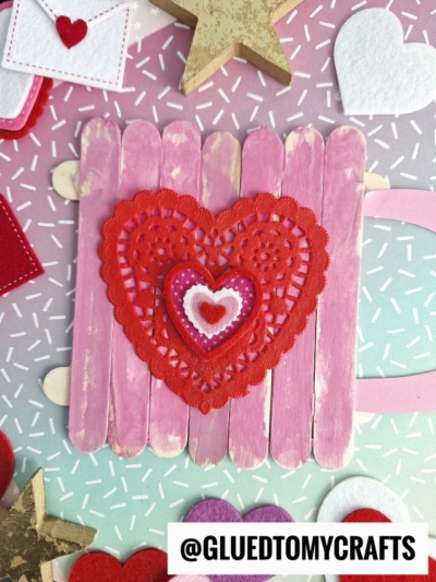 A popsicle stick cup of kindness craft in pink showcases layered hearts in red, pink, and white. The background features felt hearts, stars, and confetti-like patterns. A watermark reads @GLUEDTOMYCRAFTS.