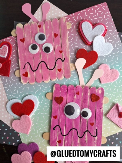 Two pink craft stick love monsters with googly eyes, heart-shaped antennae, and wavy smiles are decorated with felt hearts and placed on colorful patterned paper. @GLUEDTOMYCRAFTS is written at the bottom.