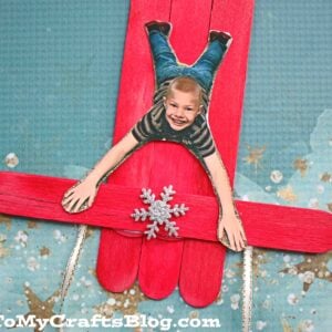 A child’s photo is cut out and placed on a popsicle stick sleigh ornament made from red sticks, glittery snowflake decor, and string runners.