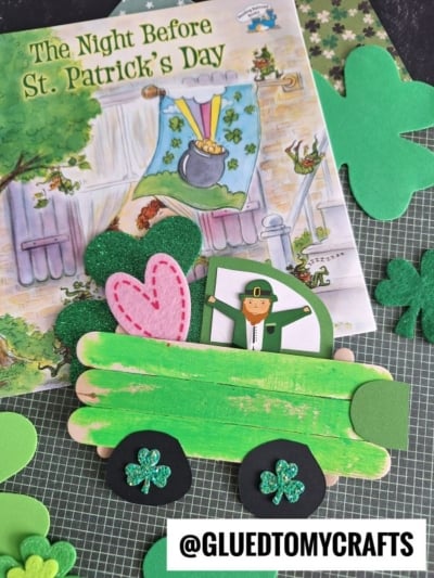 A St. Patrick’s Day-themed children’s book sits behind a popsicle stick leprechaun house, shaped like a truck and decorated with shamrocks, a paper leprechaun, and a pink heart. The Instagram handle @GLUEDTOMYCRAFTS is visible.