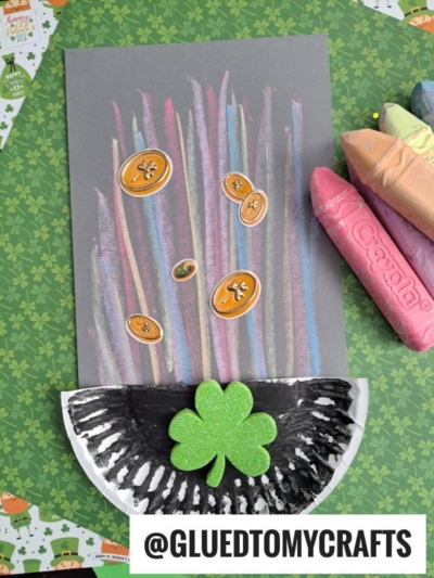 A chalk art rainbow with colorful lines and gold coins on dark paper ends in a black and white paper plate pot with a green glittery shamrock, surrounded by large Crayola crayons.