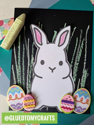 A charming bunny chalk art scene with a white bunny cutout, pink ears, and colorful Easter eggs on black paper. Chalk-drawn grass and pastel accents make this bunny art with chalk pastels truly festive! @GLUEDTOMYCRAFTS at the bottom.