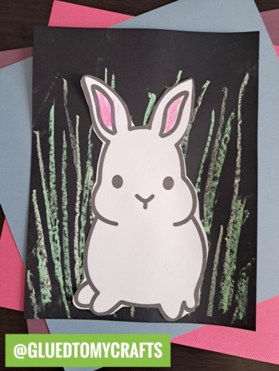 A handmade bunny cutout with pink ears is glued on black paper with crayon grass, resembling bunny chalk art. The artwork sits over blue and pink sheets, and a green label at the bottom reads @GLUETOMYCRAFTS.