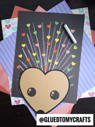 A chalk art hedgehog craft made from colored paper features a heart-shaped face and chalk-drawn quills topped by heart stickers, displayed on layered patterned paper. A piece of white chalk and the handle @GLUEDTOMYCRAFTS are visible.