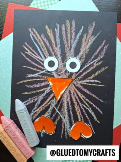 A turkey made with colorful sidewalk chalk lines on black paper, featuring large googly eyes, an orange paper beak, and orange paper feet—looking a bit like a baby bird. Pieces of pastel chalk and patterned paper sheets are in the background.
