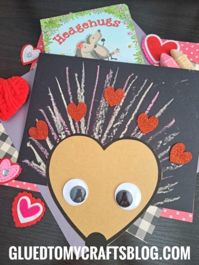 A craft project featuring a chalk art hedgehog made from a large yellow heart, googly eyes, glittery red heart shapes for spikes, and painted branches on black paper, with a Hedgehugs book and felt hearts in the background.