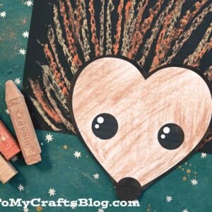 A charming chalk art hedgehog craft made with brown and peach chalk on black paper, featuring a large, heart-shaped face and big eyes, set on a teal background scattered with colorful chalk sticks.