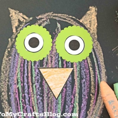 A chalk art owl craft features a drawing on black paper with colorful stripes, large green and white eyes, and a yellow beak. Four pastel crayons and a pink object sit beside the artwork on a red polka dot surface.