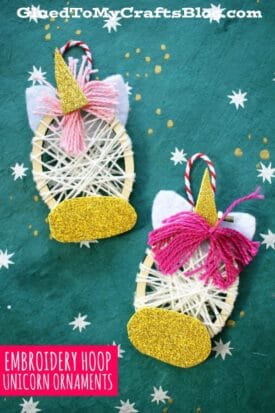 Two handmade unicorn christmas ornaments crafted from small embroidery hoops, wrapped in white yarn with pink yarn manes, gold glittery horns and snouts, and felt ears, are displayed on a green background with white star patterns.