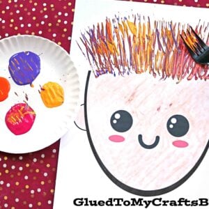 A paper with a smiling face features fork painted silly hair. Beside it, a paper plate holds purple, orange, pink, and yellow paint, with a plastic fork nearby—all set on a cheerful red polka-dotted background.