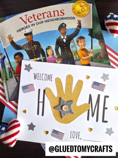 A children’s book titled Veterans: Heroes in Our Neighborhood sits behind a handmade welcome home card featuring a yellow handprint keepsake. The card is decorated with stars, hearts, and small American flags.