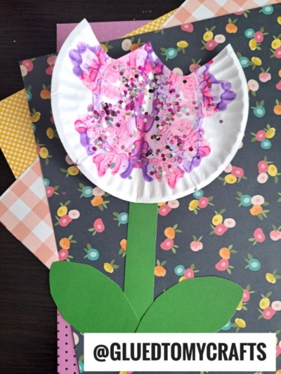 A handmade paint splat flower craft with a colorful, painted coffee filter blossom, decorated with glitter and a green paper stem and leaves, placed on floral-patterned paper. A watermark reads @GLUEDTOMYCRAFTS.