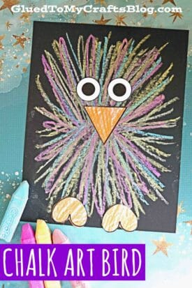 A colorful sidewalk chalk baby bird drawing made with chalk on black paper, featuring large cartoon eyes, an orange beak, and orange feet. Colored chalk sticks lie nearby. The caption at the bottom reads CHALK ART BIRD.