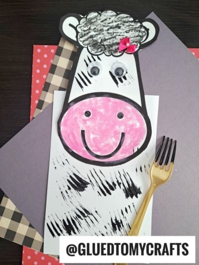 A handmade cow craft made from paper, with a pink face, googly eyes, black spots, a flower on its head, and a fork painted zebra-style nearby. Several colored papers are layered underneath. Text reads @GLUEDTOMYCRAFTS.