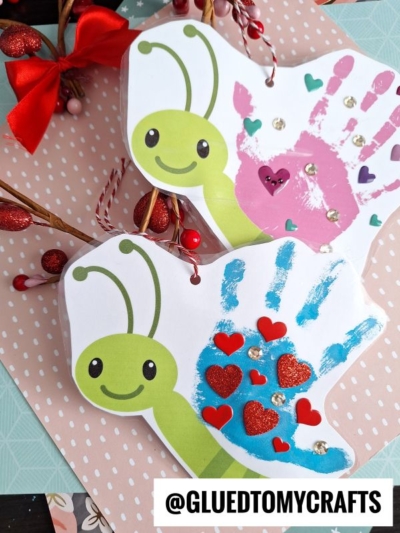 Two butterfly crafts made from paper and painted handprints shine as a butterfly handprint Mother's Day card, each with a smiling green head and colorful wings decorated with heart and jewel stickers. A festive background with bows and berries adds charm.