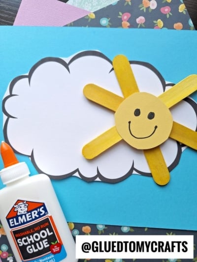 A cheerful popsicle stick sunshine with a smiling face peeks from behind a white cloud on blue paper. Elmer's school glue and patterned papers sit nearby. @GLUEDTOMYCRAFTS is written at the bottom.