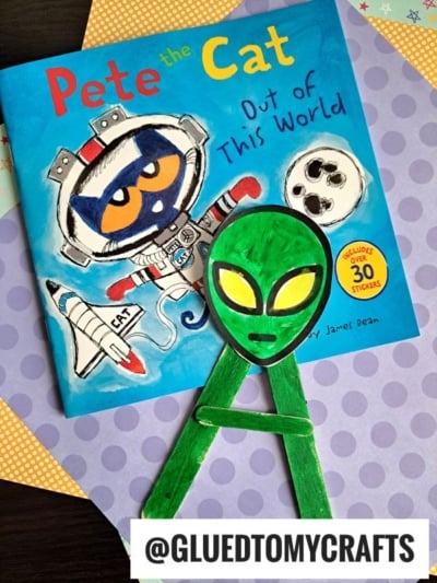 A Pete the Cat: Out of This World book sits on purple star-patterned paper, next to a popsicle stick alien made from craft sticks. An @GLUEDTOMYCRAFTS label is at the bottom.